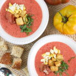 Two bowls of soup are placed next to croutons and a tomato.