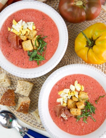 Two bowls of soup are placed next to croutons and a tomato.