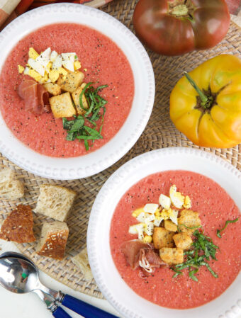 Two bowls of soup are placed next to croutons and a tomato.