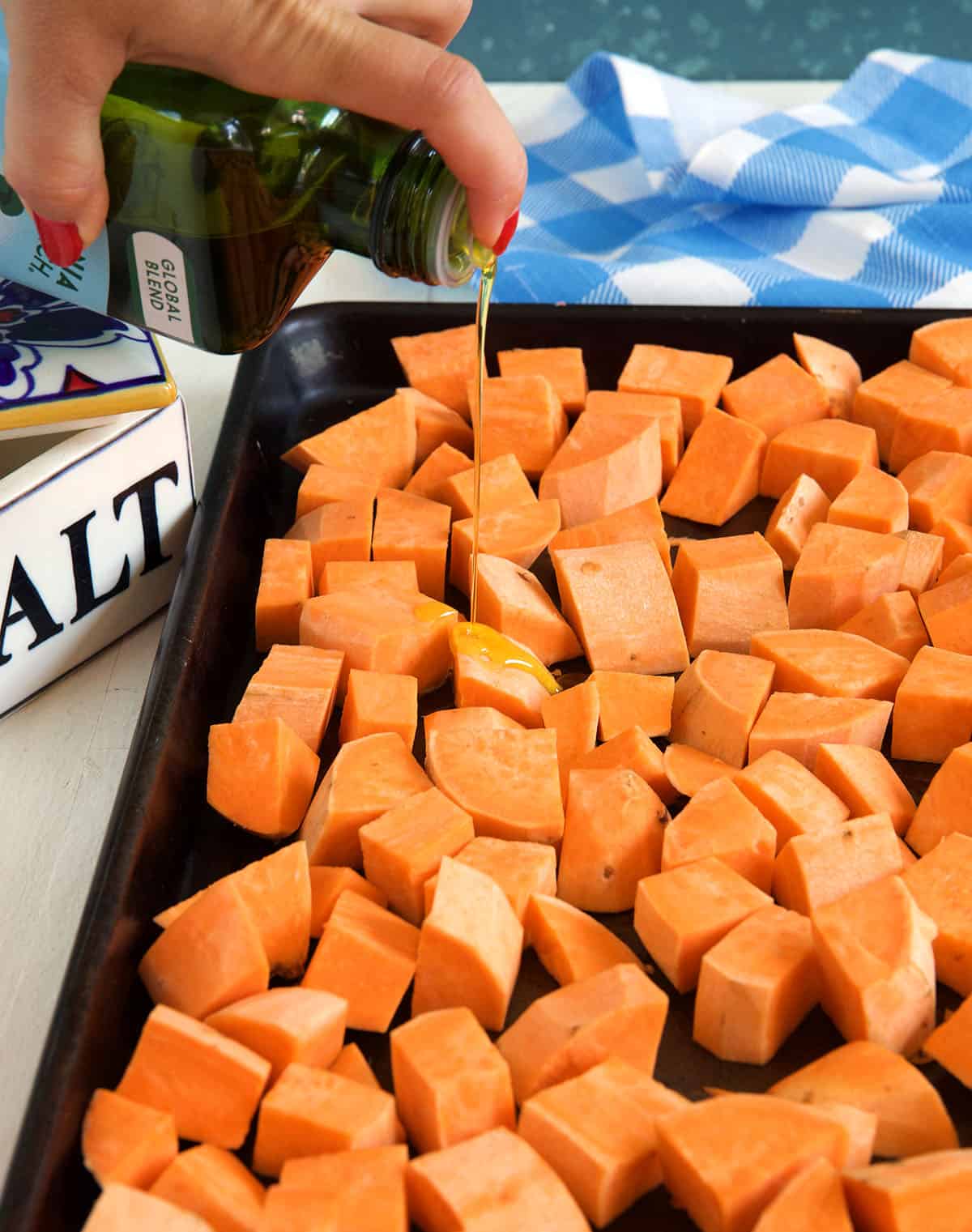 Olive oil is being drizzled onto a sheet pan full of chopped sweet potatoes. 