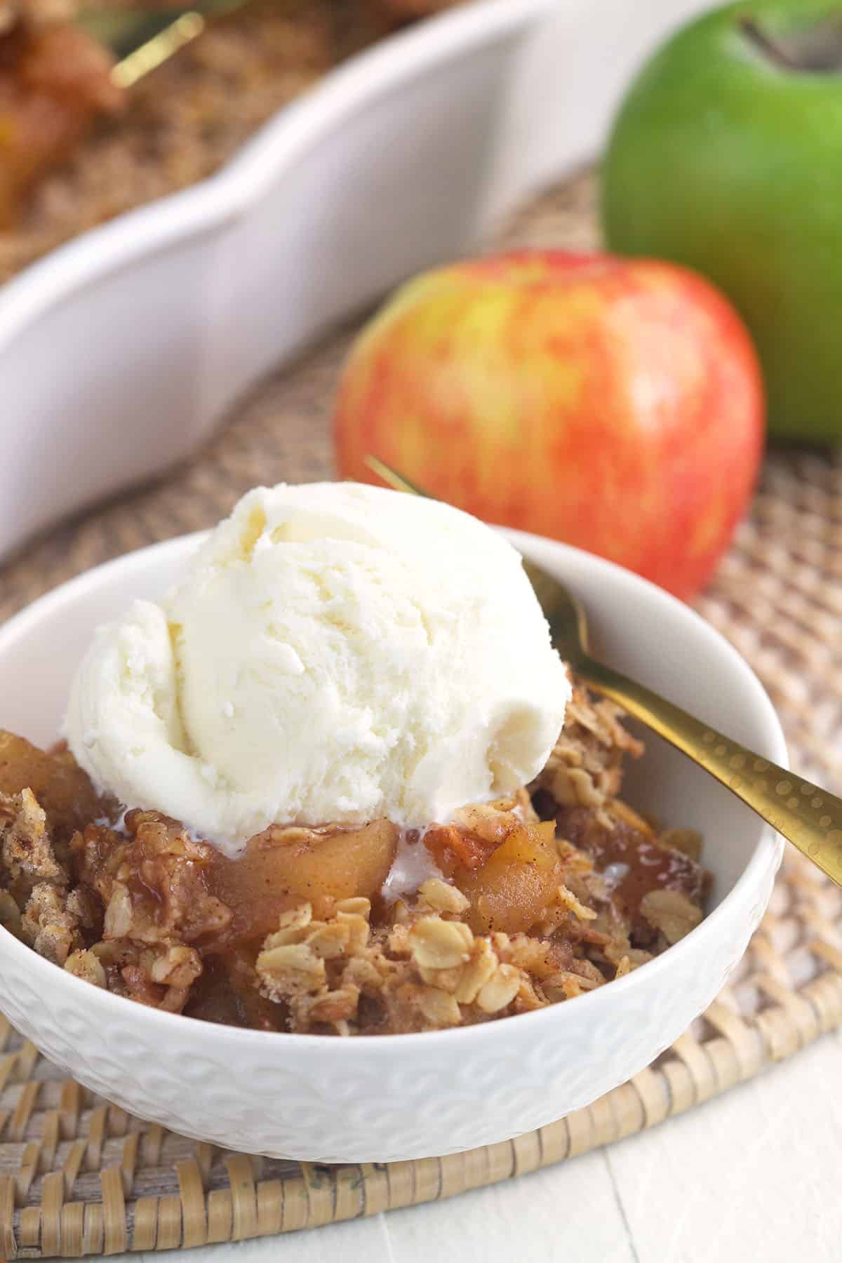 Apples are placed behind a bowl filled with apple crisp and ice cream. 