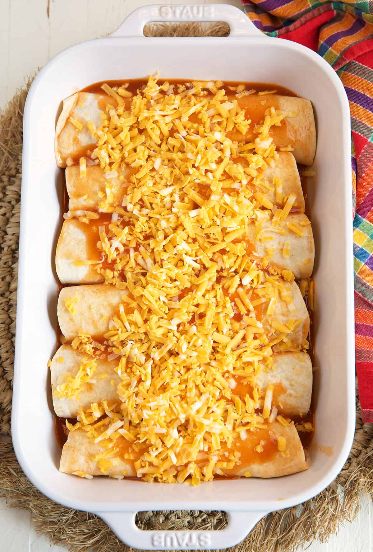 Shredded cheese and enchilada sauce are spread all over uncooked enchiladas in a baking dish.