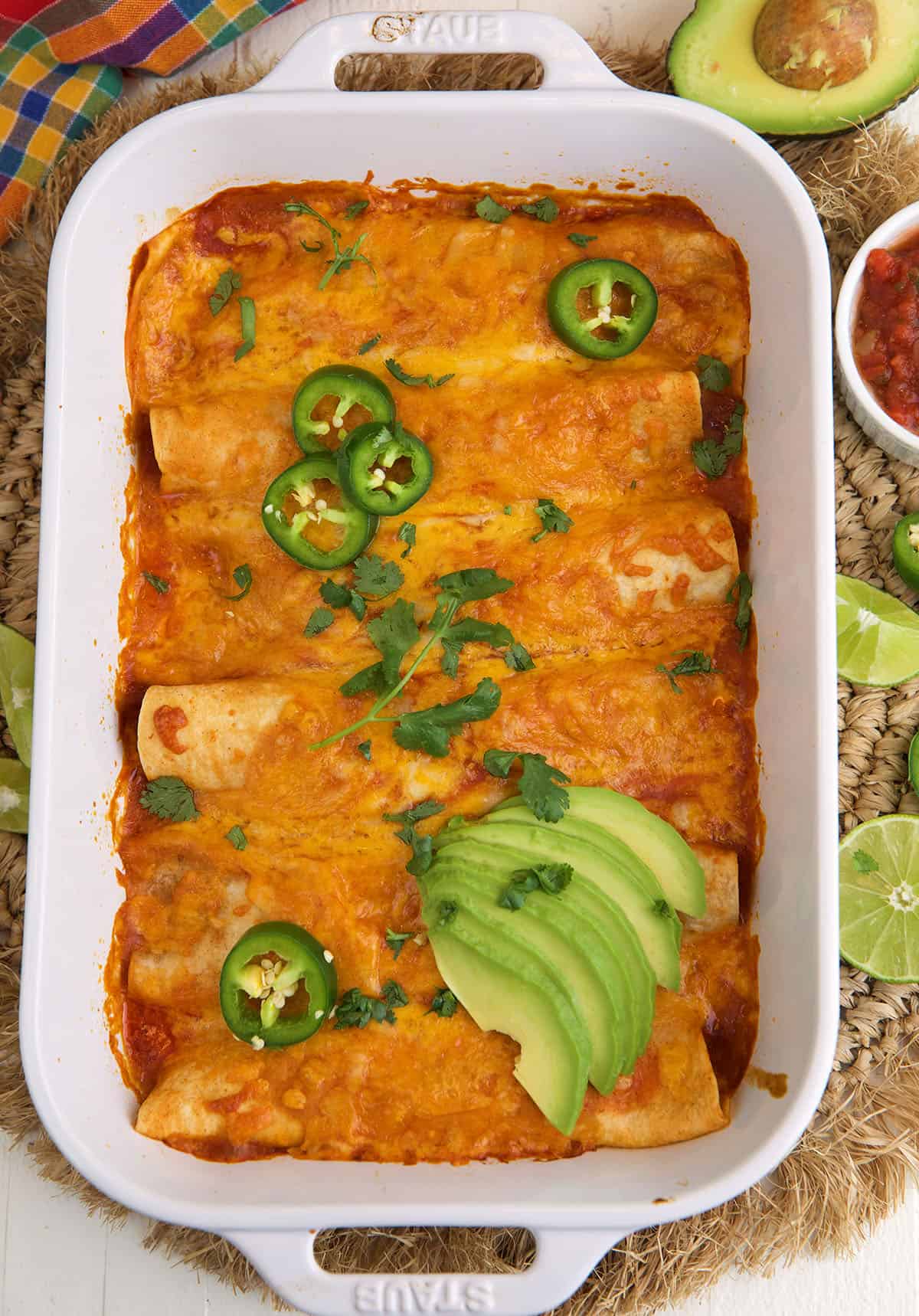 A baking dish is filled with cooked beef enchiladas and is garnished with avocado and jalapenos. 