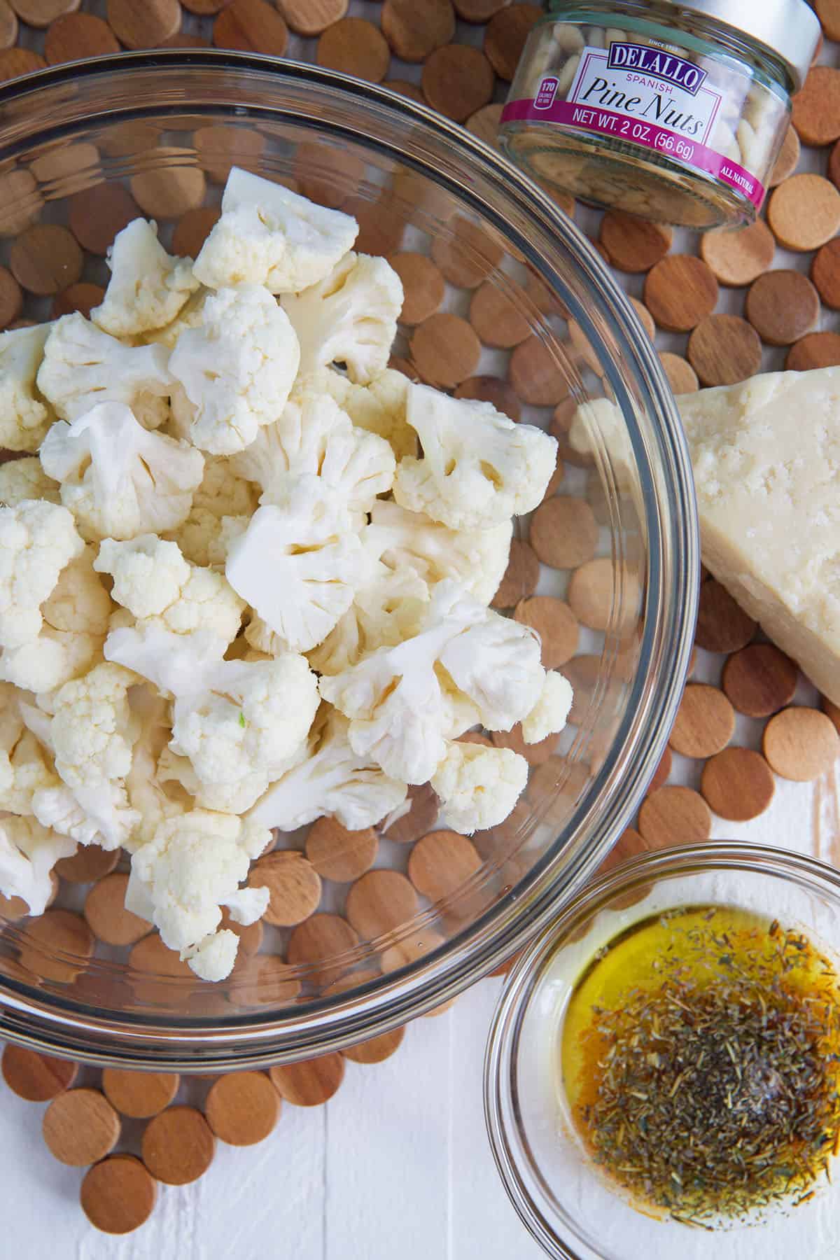 A glass bowl filled with raw cauliflower is placed next to seasoned olive oil and pine nuts. 