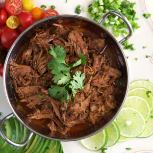 Limes, onions and tomatoes are placed around a small pot filled with barbacoa.
