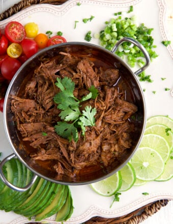 Limes, onions and tomatoes are placed around a small pot filled with barbacoa.