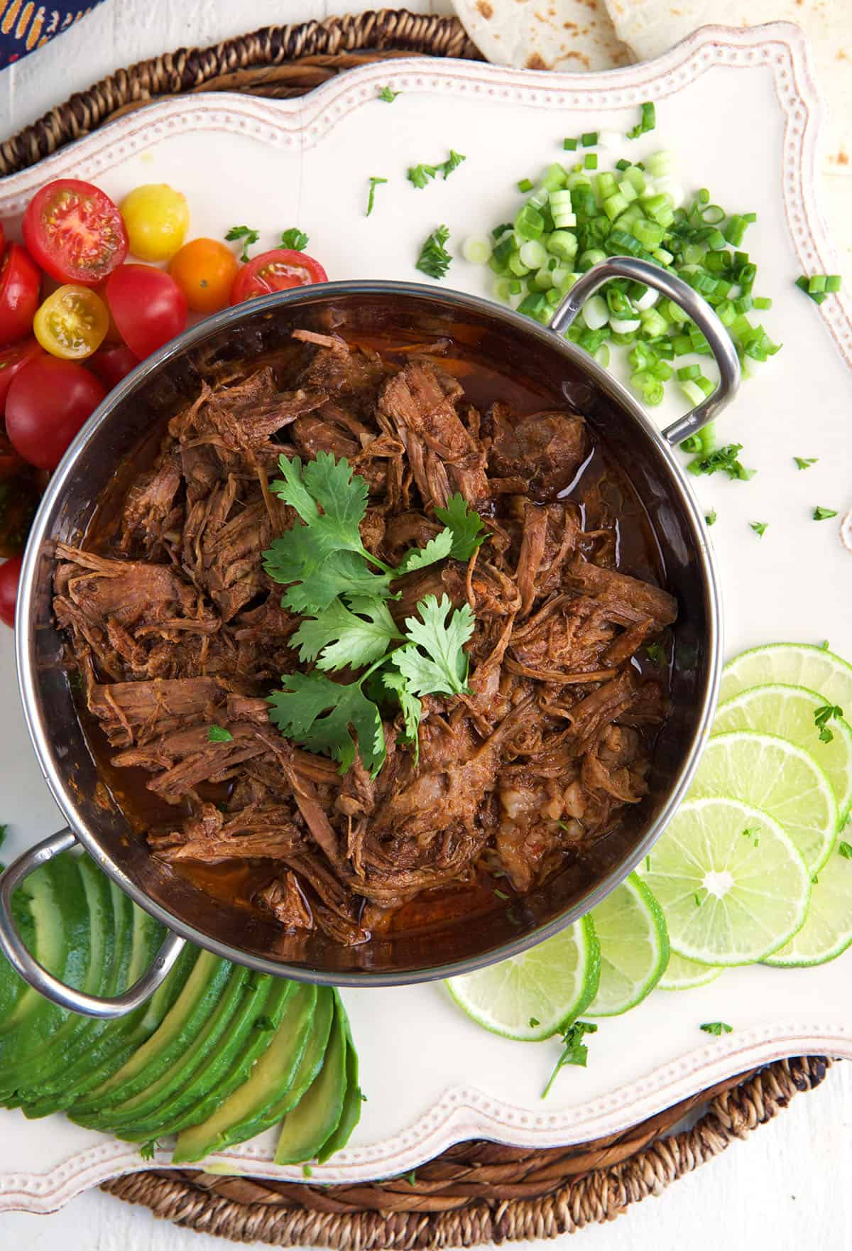 Limes, onions and tomatoes are placed around a small pot filled with barbacoa. 
