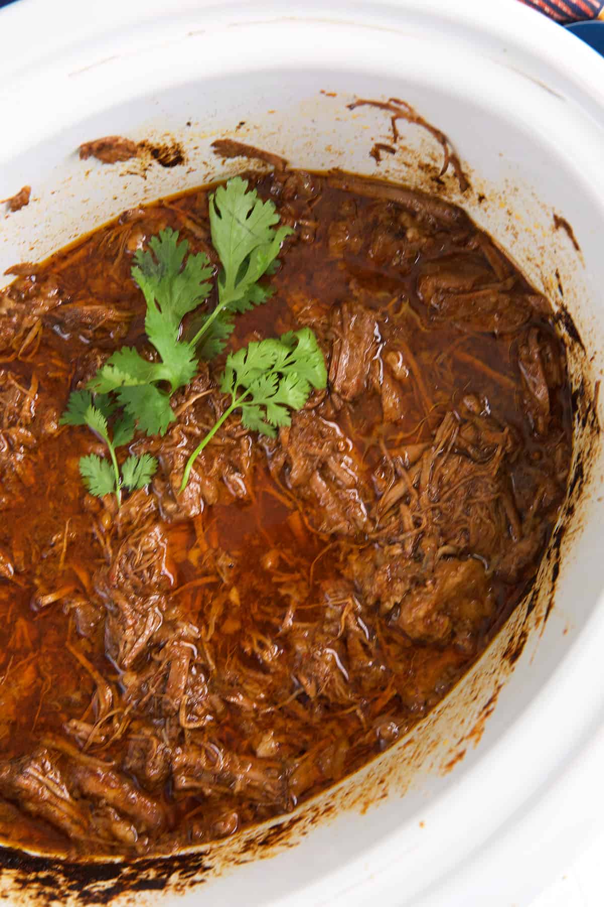 A white Crockpot is filled with cooked beef barbacoa. 