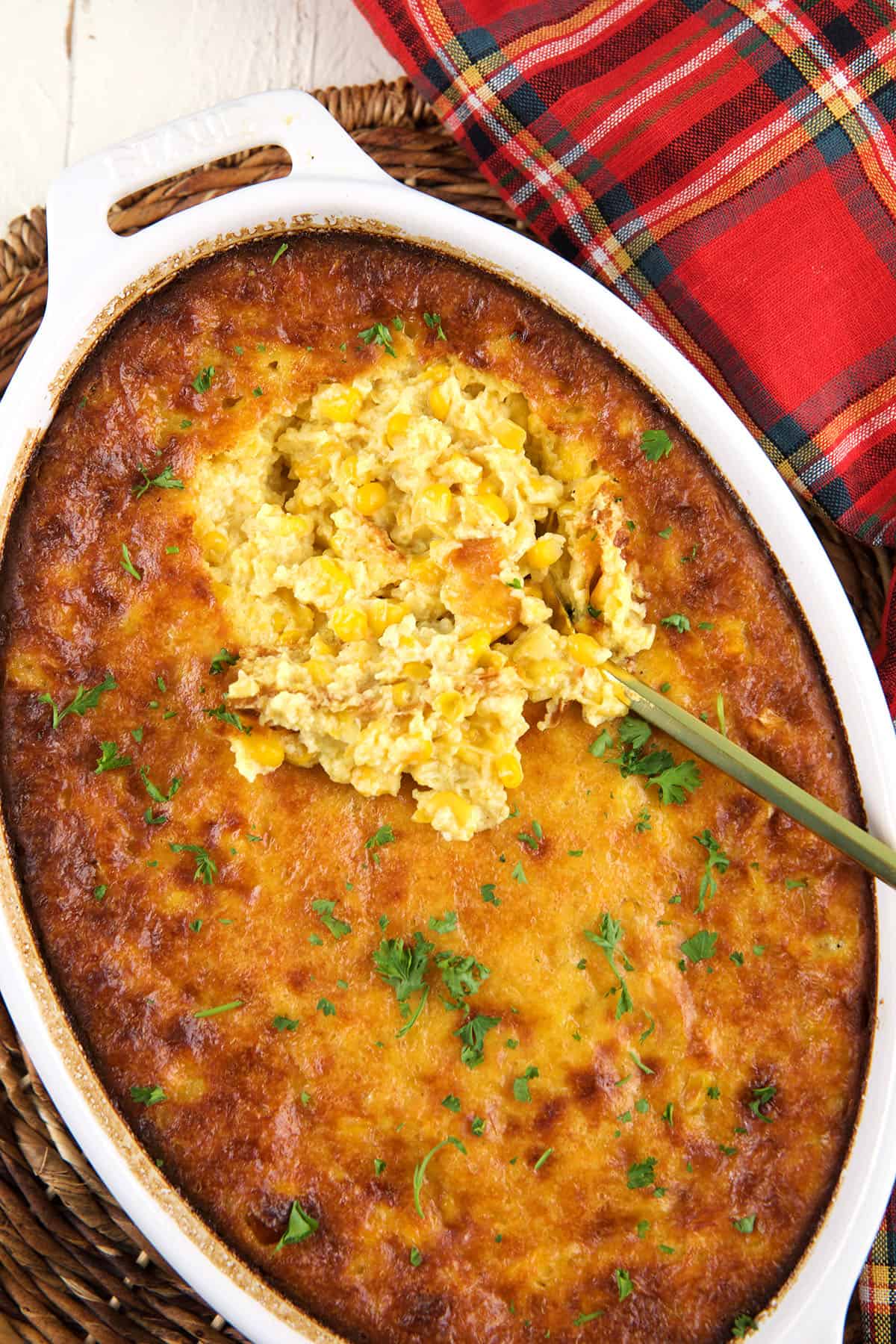 A white ovular baking dish is filled with corn pudding beneath a full scoop. 