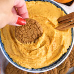 A hand is dipping a cookie into a garnished bowl of pumpkin dip.