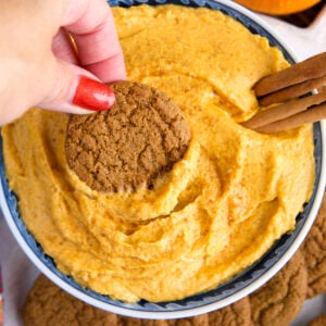 A hand is dipping a cookie into a garnished bowl of pumpkin dip.