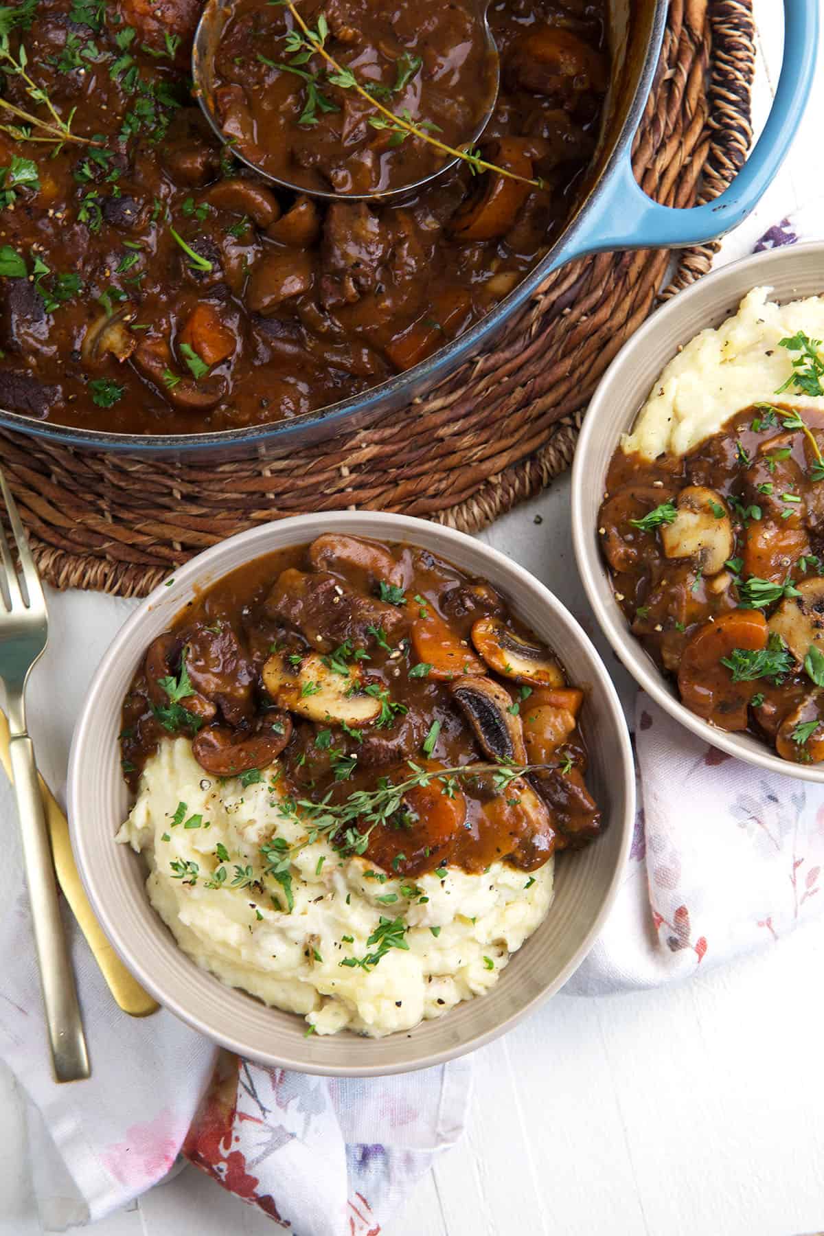Two bowls of beef bourguignon are placed next to a full skillet. 