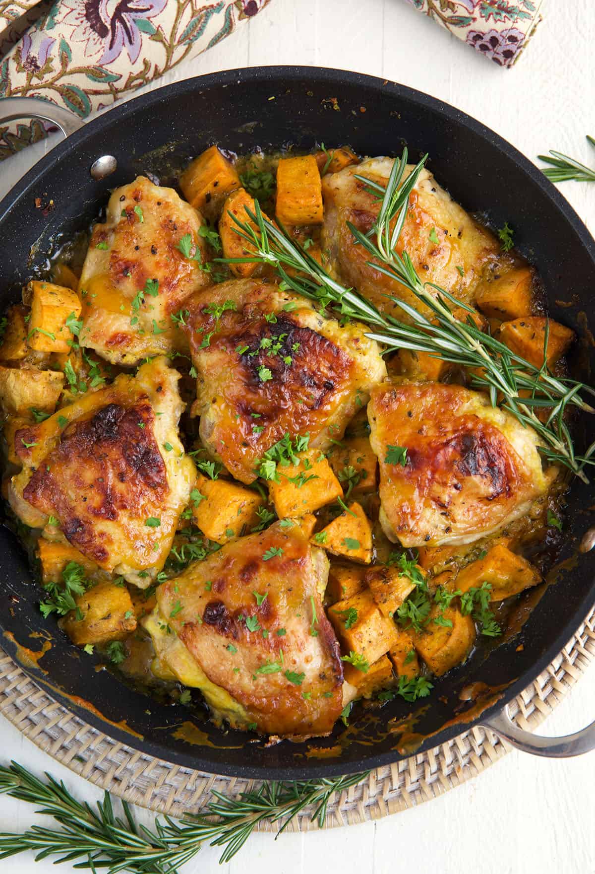 A black skillet is filled with herbs, chicken thighs and sweet potatoes.