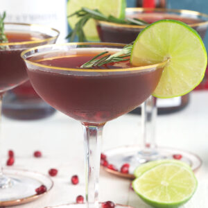 A pomegranate martini is presented next to scattered seeds and limes.