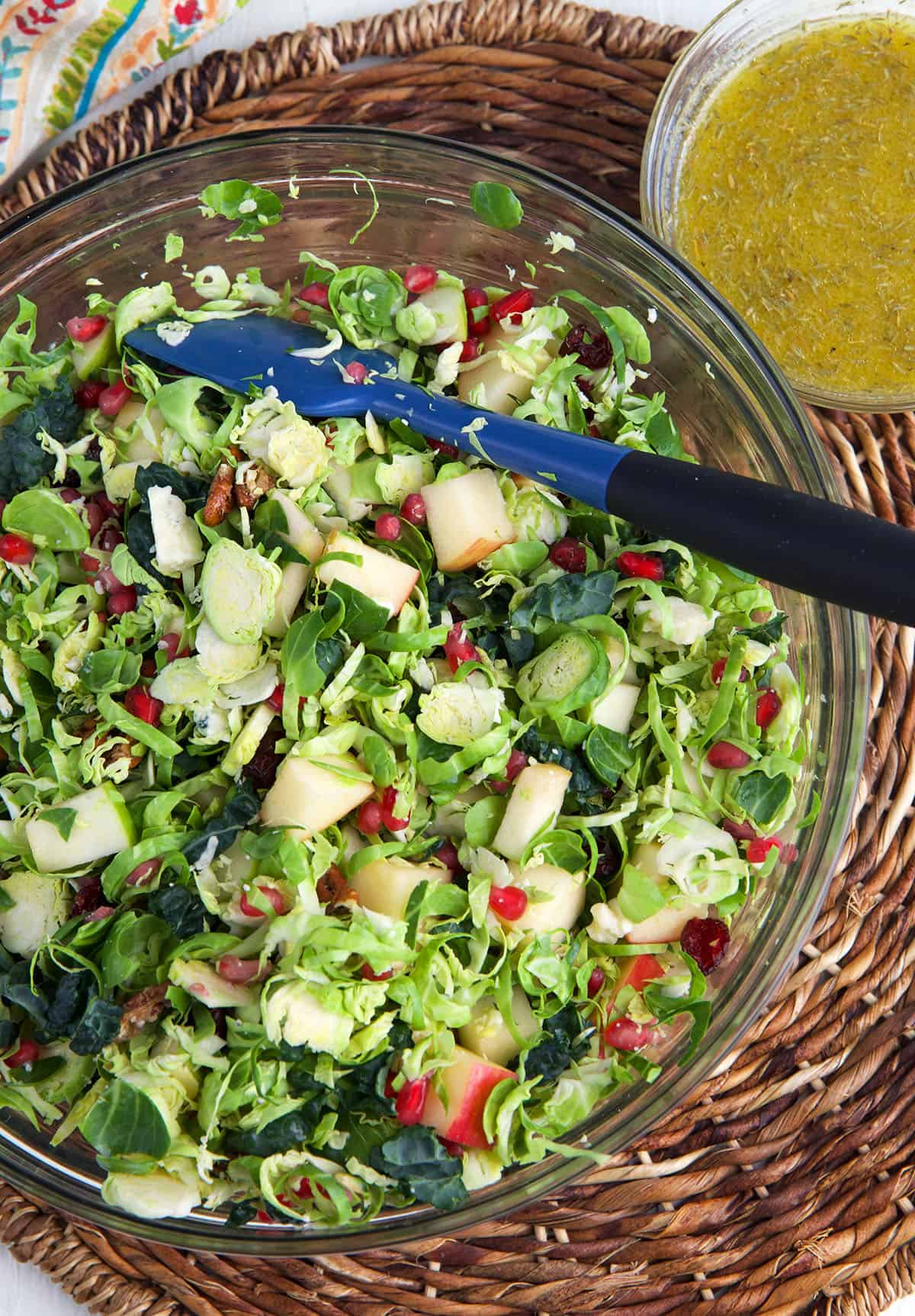 A brussel sprout salad has been tossed in a large bowl and is placed next to a small bowl of vinaigrette. 