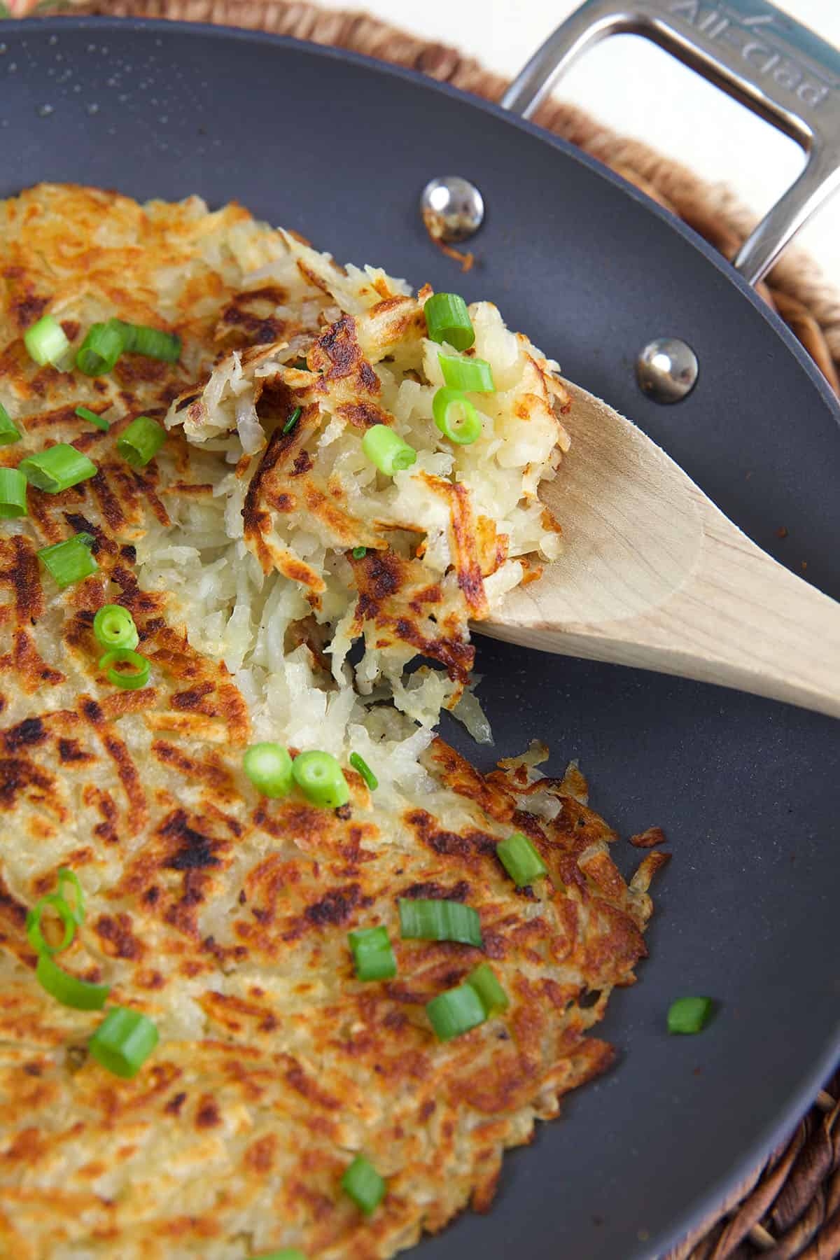 A wooden spoon is lifting a small portion of hash browns from the skillet. 