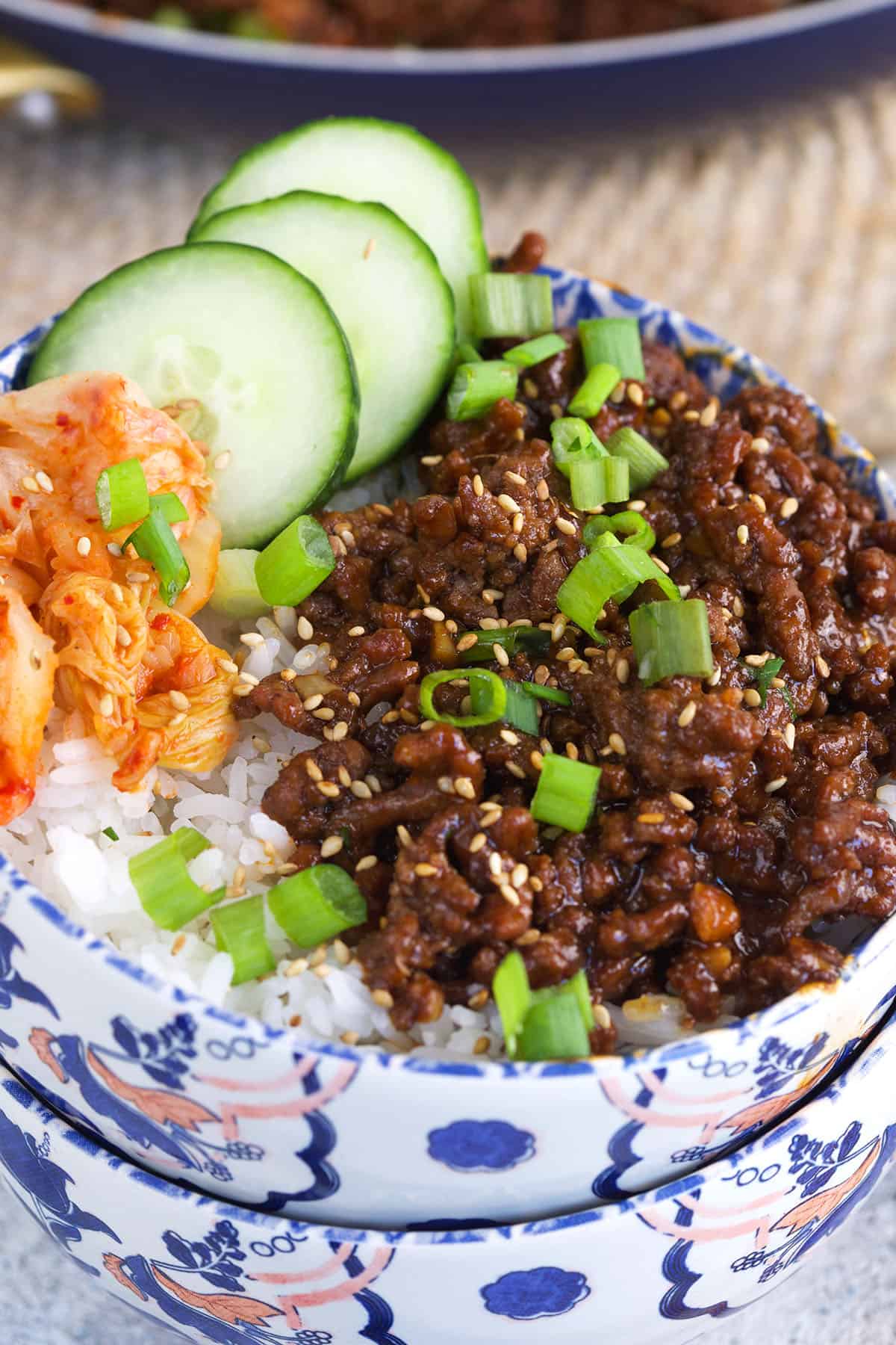 Cucumbers, kimchi and green onions are placed in a bowl with rice and ground beef. 