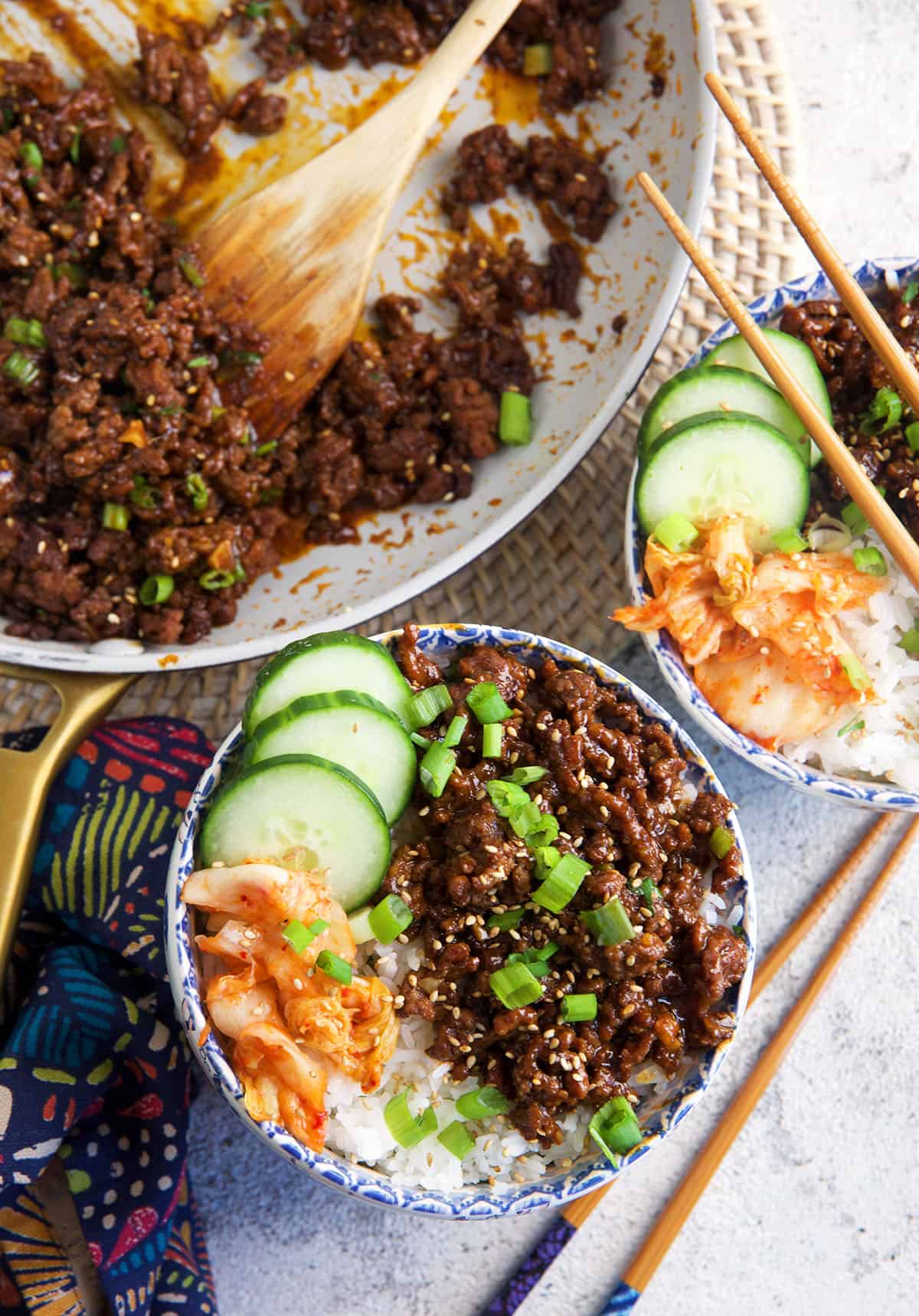 Two bowls of Korean ground beef and veggies are placed next to a skillet. 