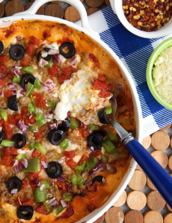 Pizza dip topped with olives, peppers, sausage and pepperoni with a serving spoon in it.