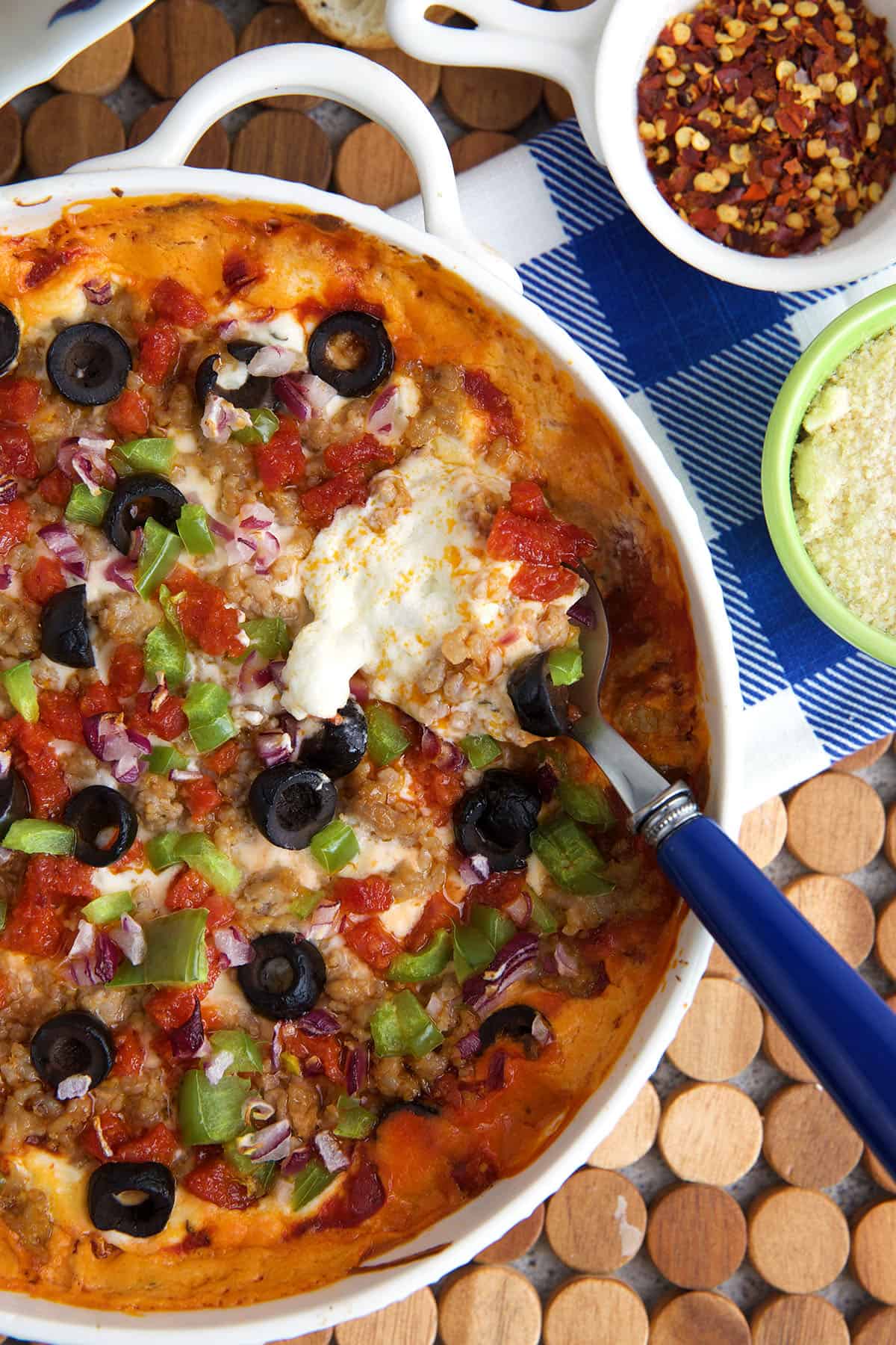 Pizza dip topped with olives, peppers, sausage and pepperoni with a serving spoon in it.