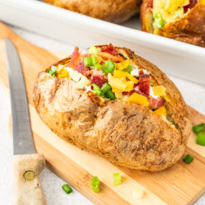 A baked potato is fully garnished and placed on a wooden cutting board.