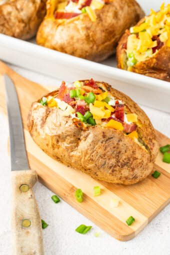 A baked potato is fully garnished and placed on a wooden cutting board.
