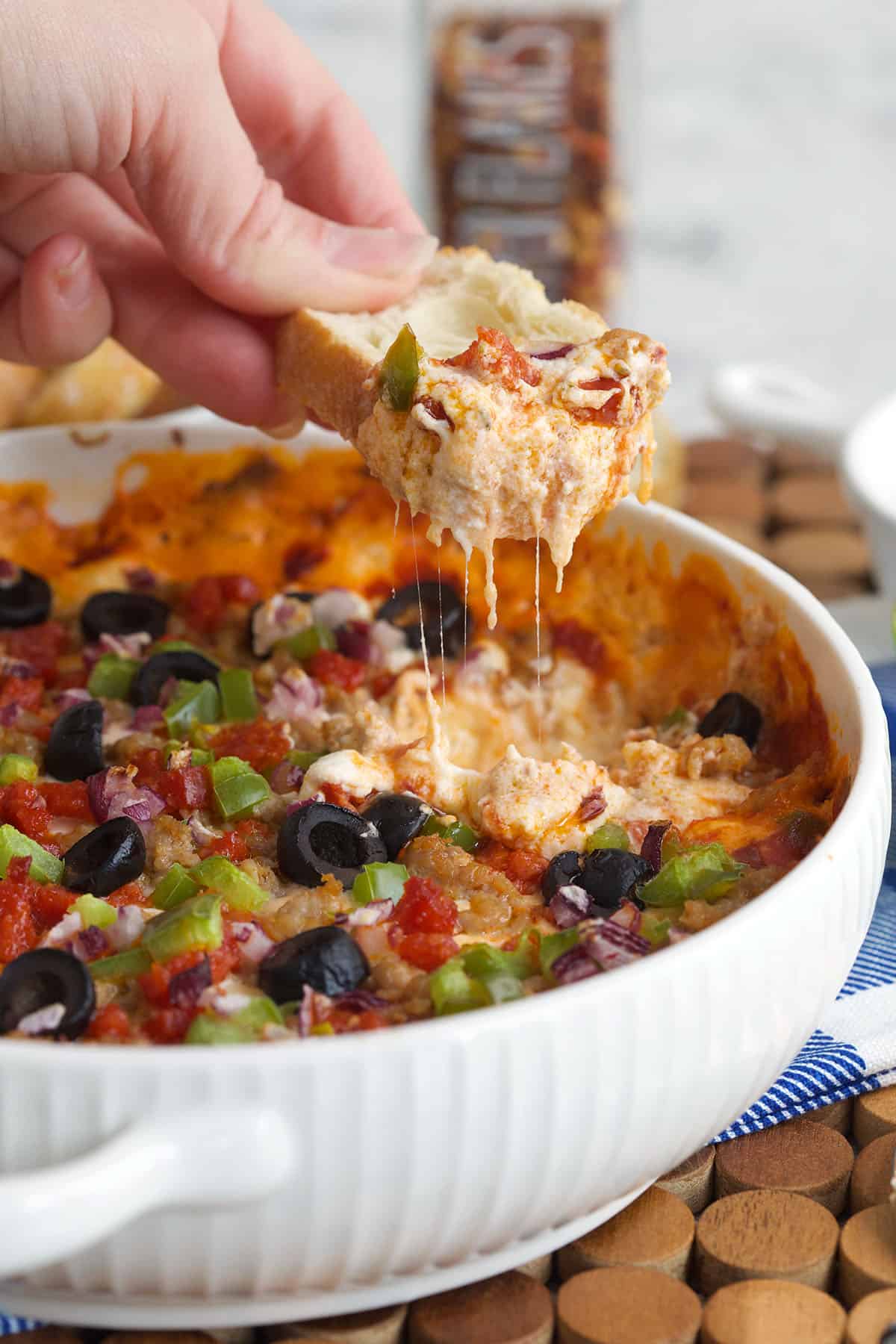 Pizza dip in a white casserole with a hand dipping a crostini into it with a cheese pull into the dish.