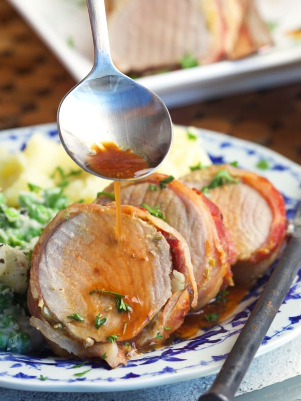 Juices are being drizzled over a cooked pork tenderloin wrapped in bacon.