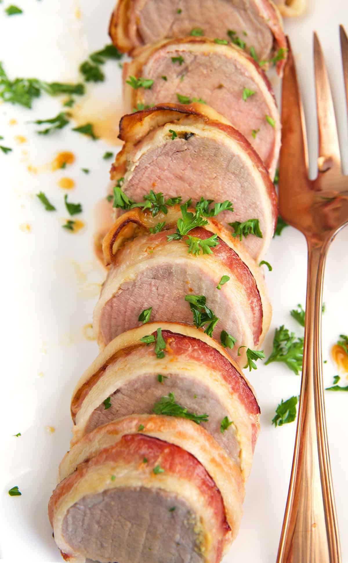 Parsley is sprinkled on top of a bacon wrapped pork tenderloin. 