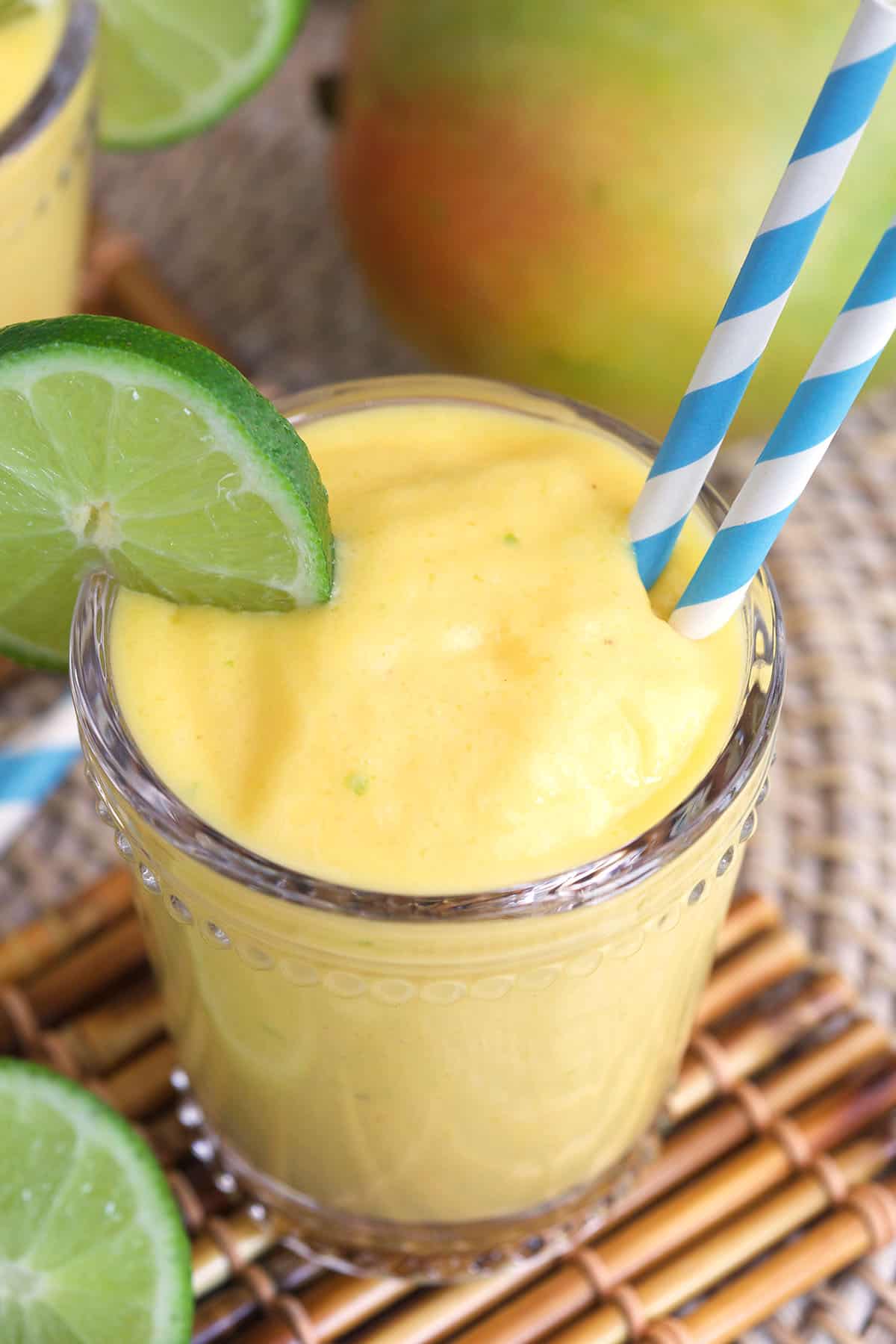 Creamy Mango Smoothie in a glass with a lime slice on the side and two blue and white striped straws. Glass is placed on top of a bamboo coaster.