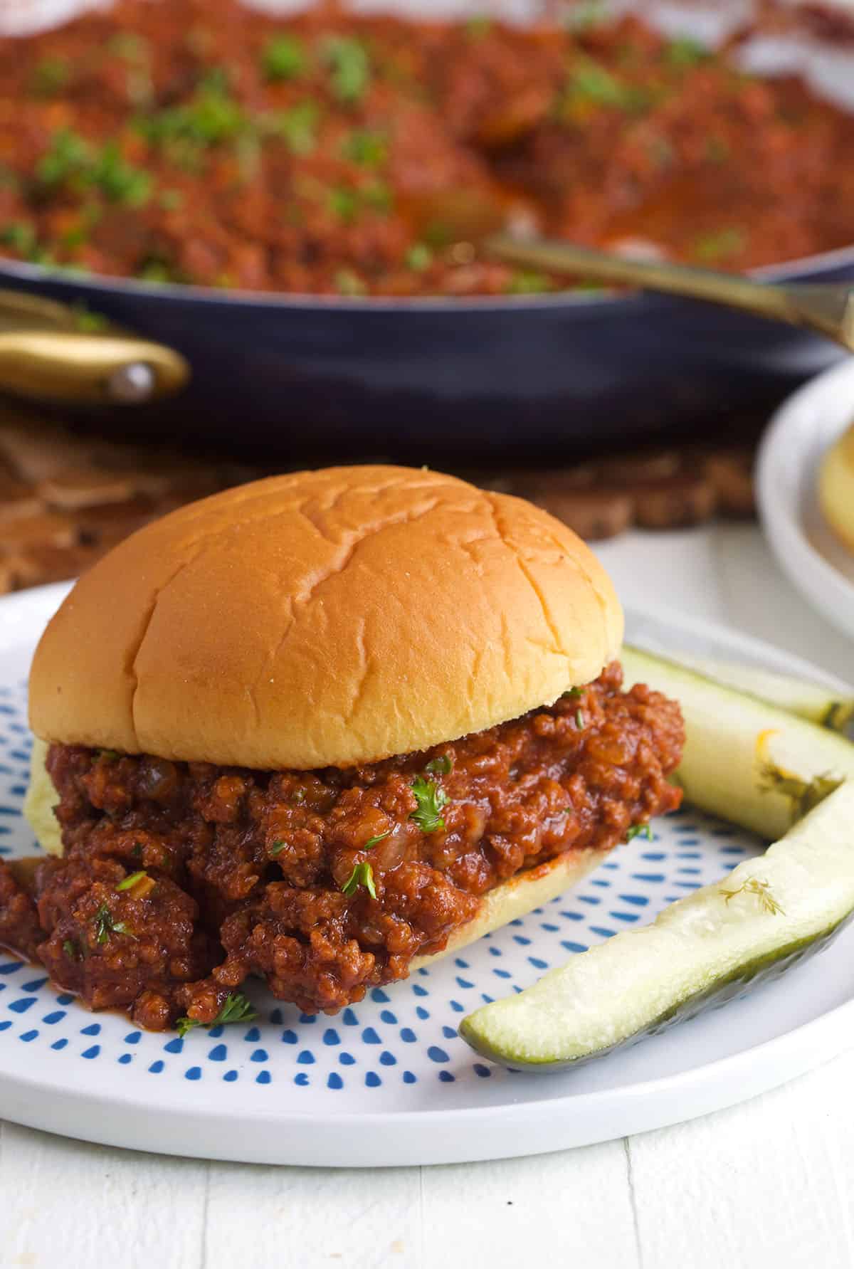 Sloppy Joe on a bun with pickles on a white plate with blue spots.