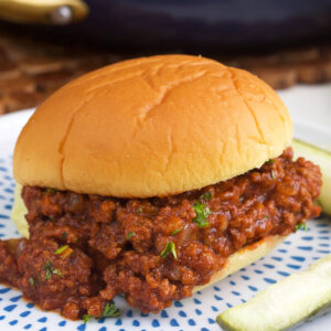 Sloppy Joe on a potato roll with a pickle on a plate.