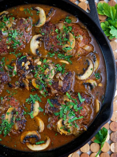 Salisbury Steak recipe in a cast iron skillet with mushroom gravy and sprinkled with chopped parsley.