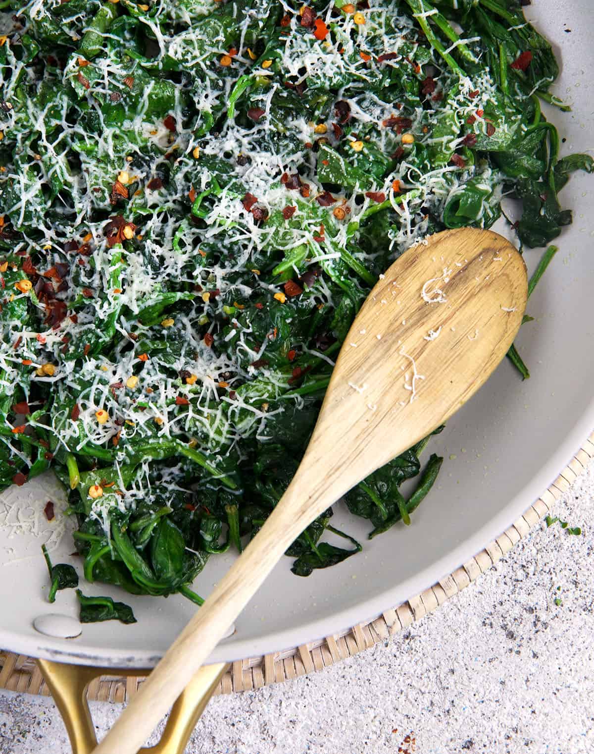 A wooden spoon is placed in a skillet with sauteed spinach. 