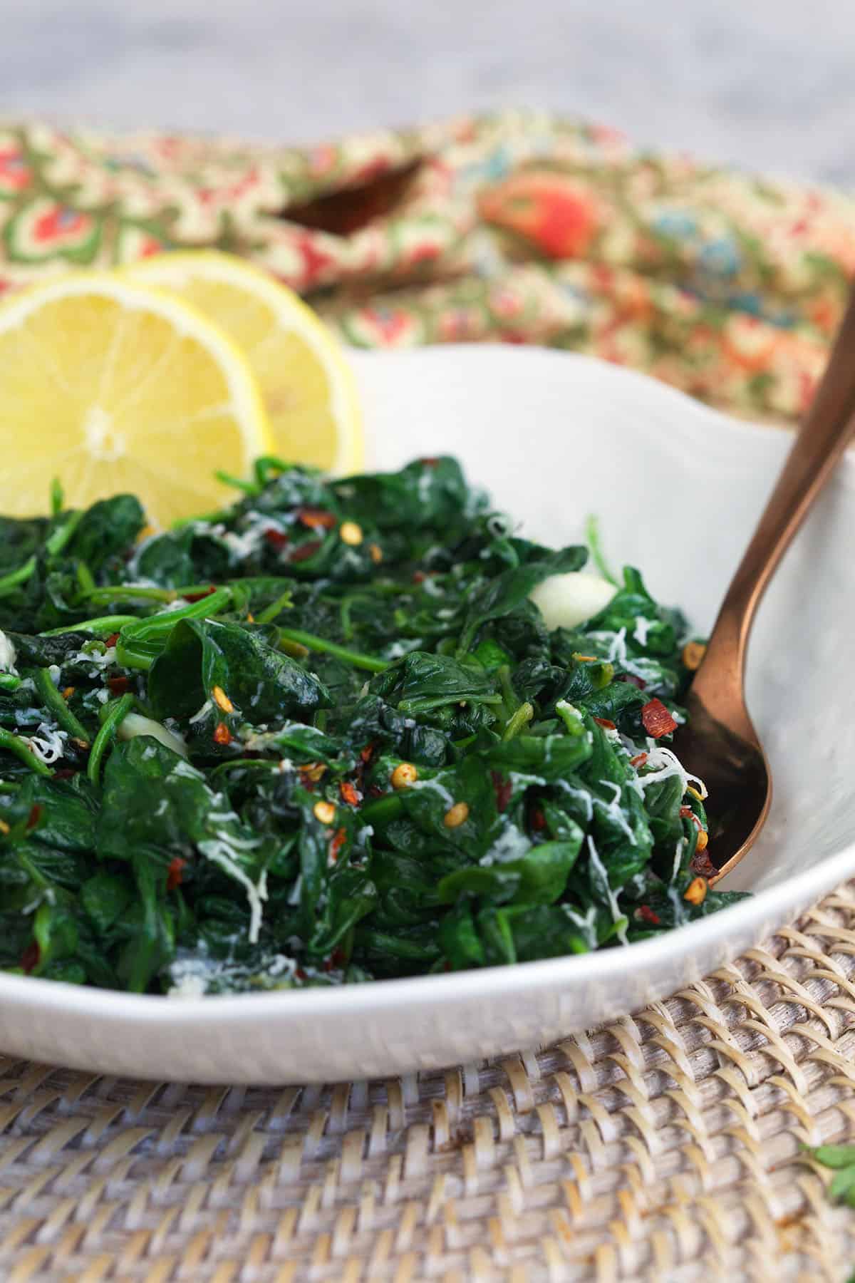 Lemon slices are placed in a white bowl filled with cooked spinach. 