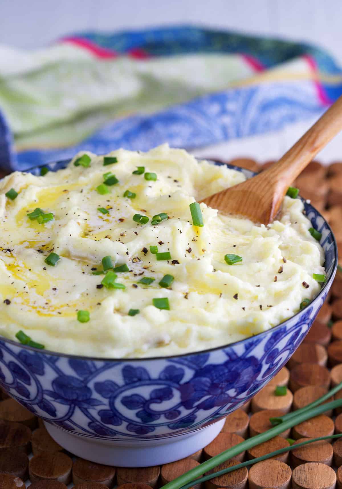 Mashed potatoes in a blue and white bowl topped with chives and butter with a wooden spoon dug inside.