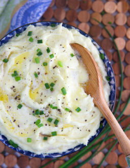 Mashed Potatoes in a blue and white bowl topped with butter and chives with a wooden spoon in them.