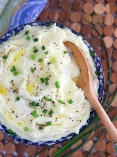 Mashed Potatoes in a blue and white bowl topped with butter and chives with a wooden spoon in them.