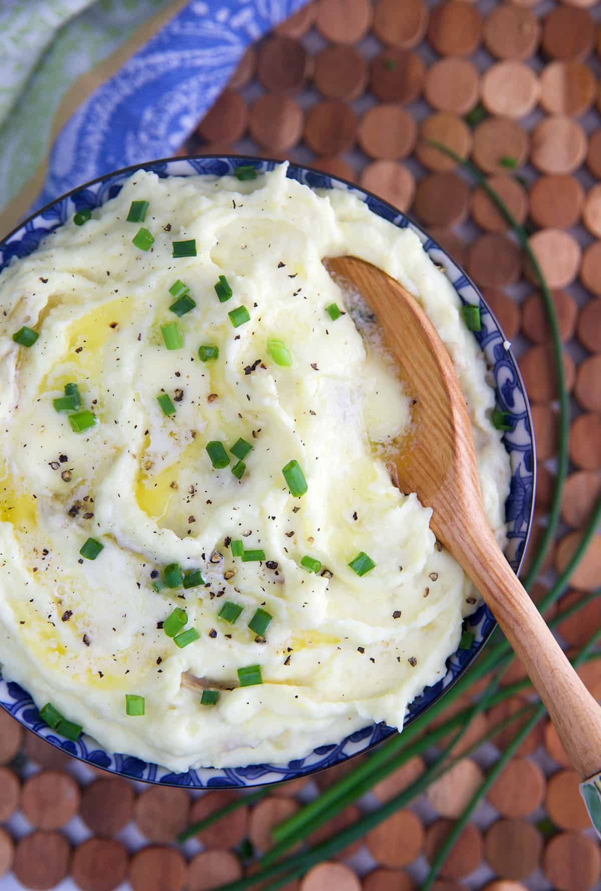 Mashed Potatoes in a blue and white bowl topped with butter and chives with a wooden spoon in them.