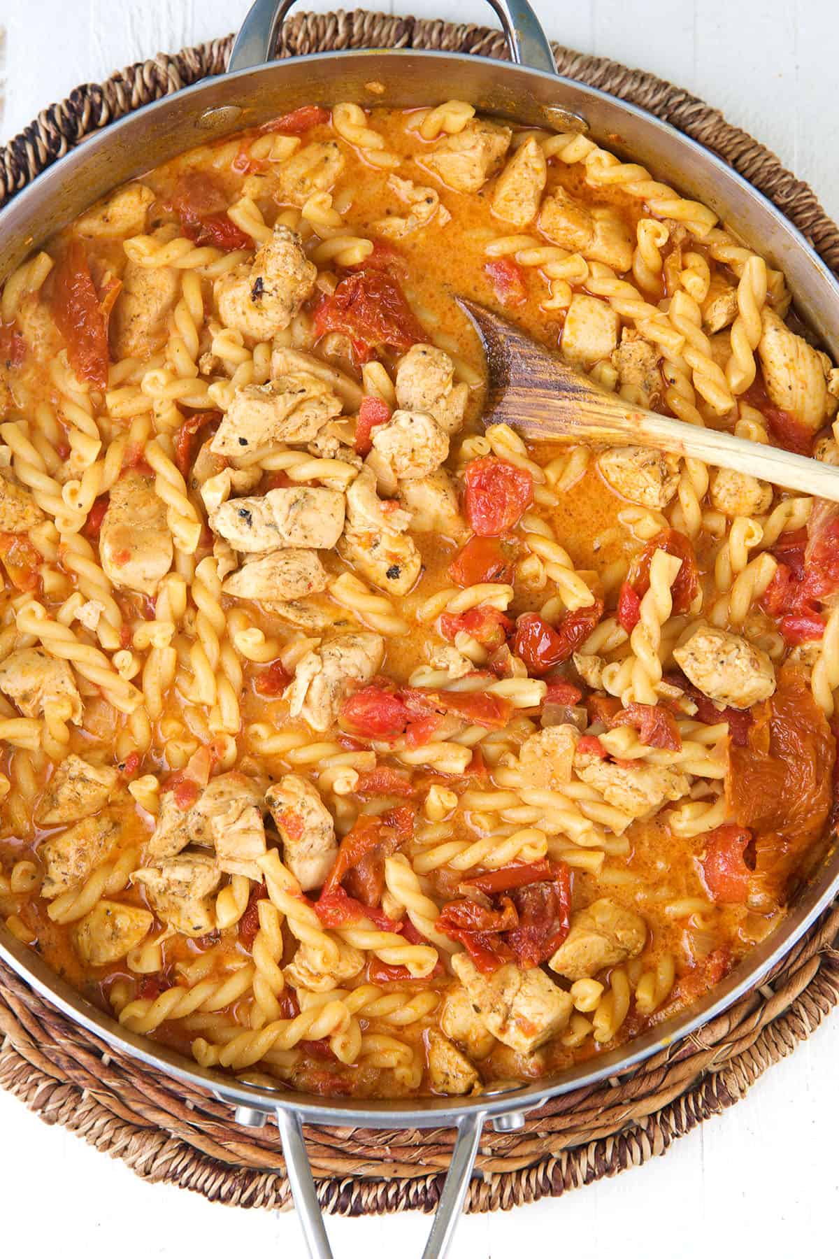 A wooden spoon is placed in a skillet filled with chicken pasta. 