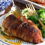 A bacon wrapped chicken breast is being drizzled with pan drippings.