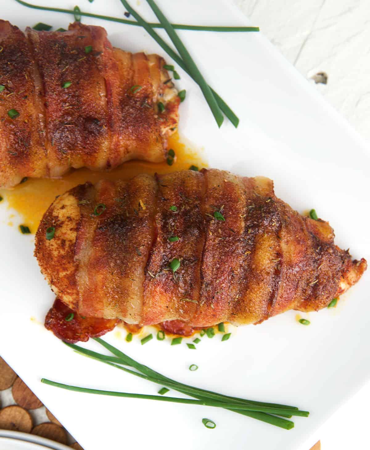Cooked chicken breasts wrapped in bacon are on a white plate with herbs. 