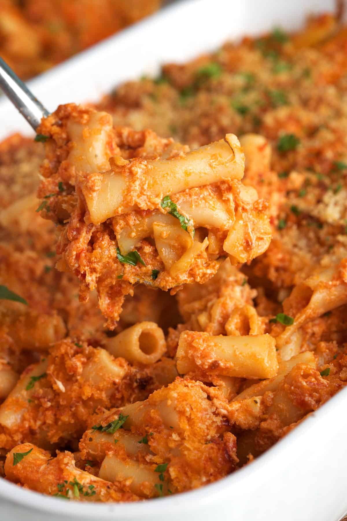 A large serving spoon is lifting a portion of ziti from the baking dish. 