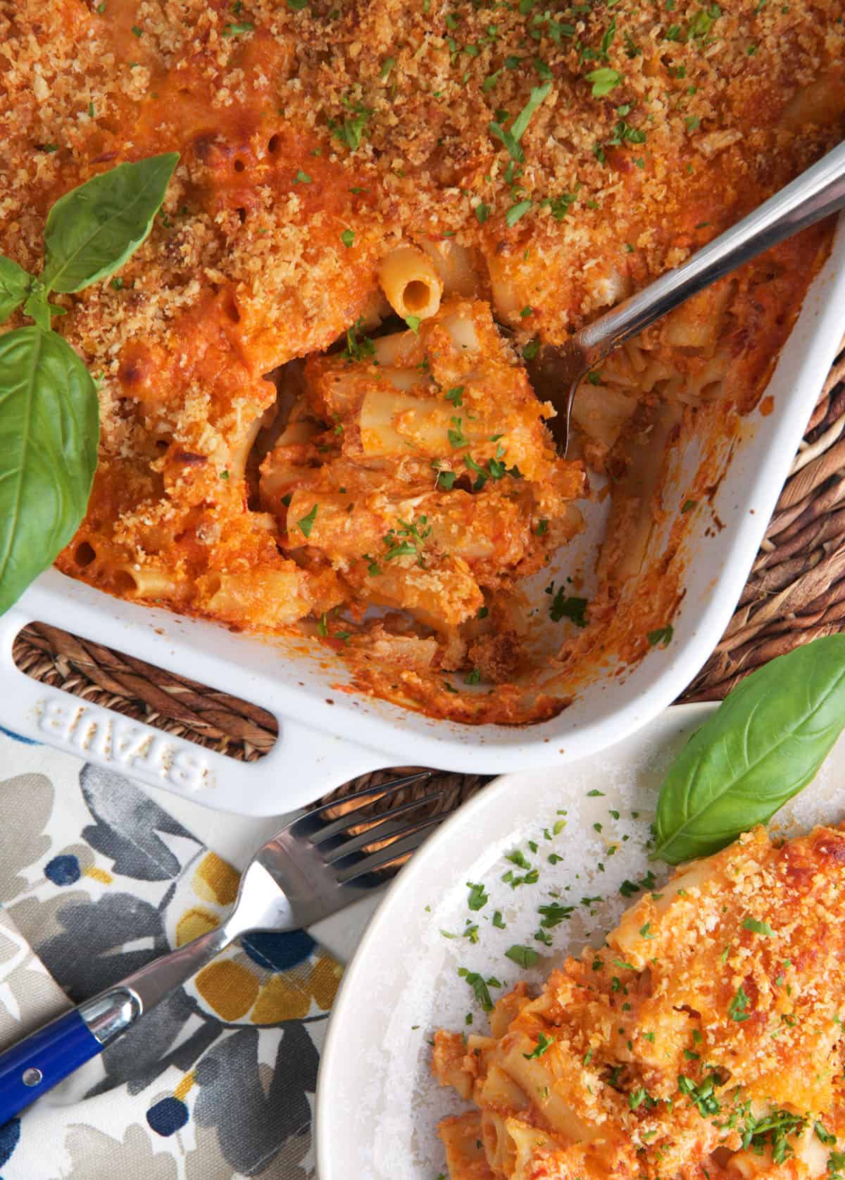 A plate is placed next to a casserole dish filled with baked ziti. 