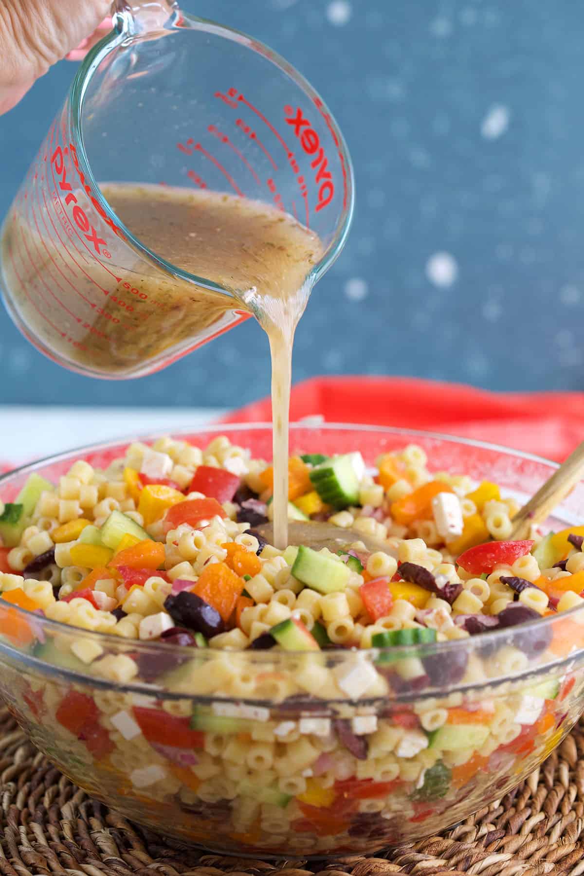 Greek Salad Dressing being poured over pasta salad in a glass bowl.