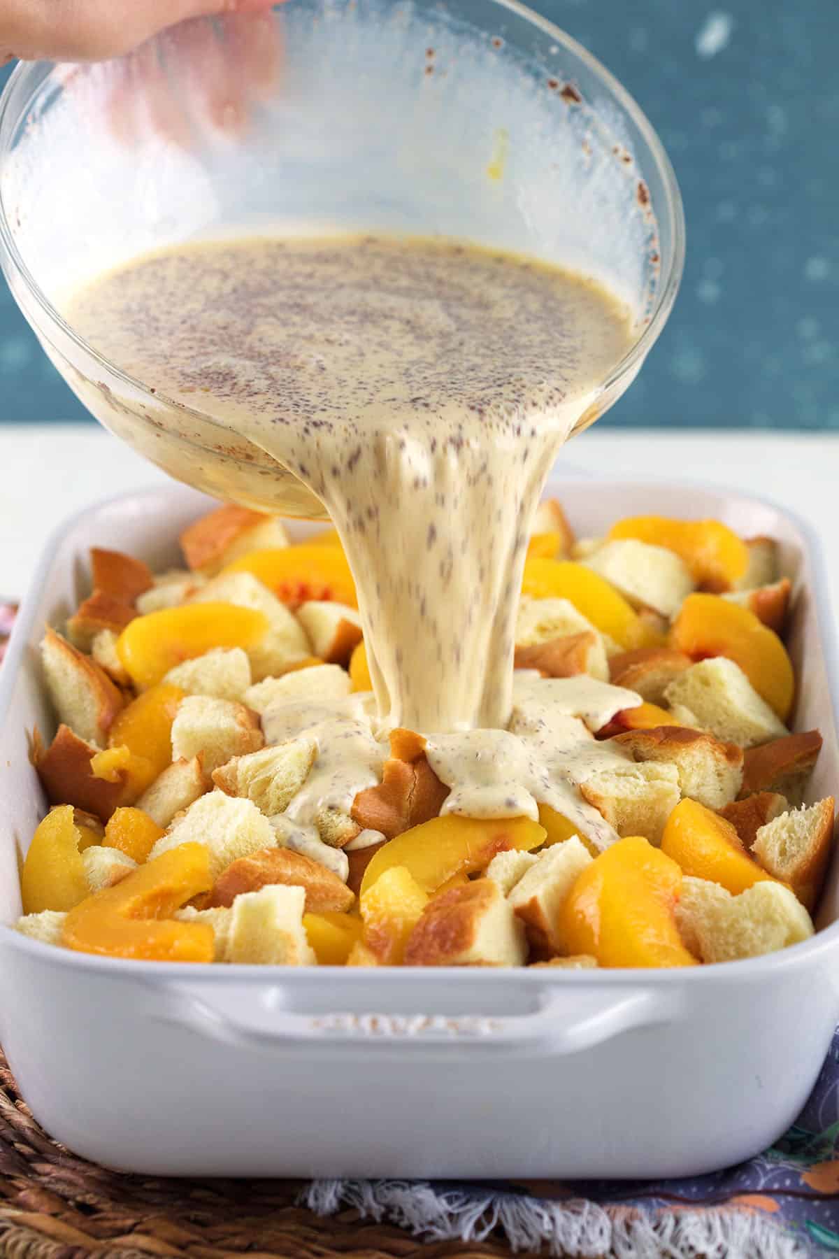 An egg mixture is being poured on top of peaches and bread in a baking dish. 