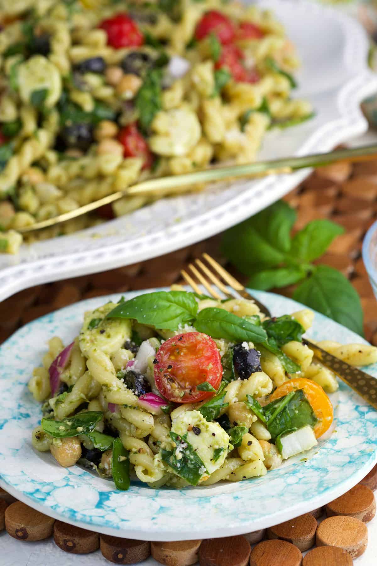 A plate of pasta salad is placed next to a full sized platter of pasta salad. 