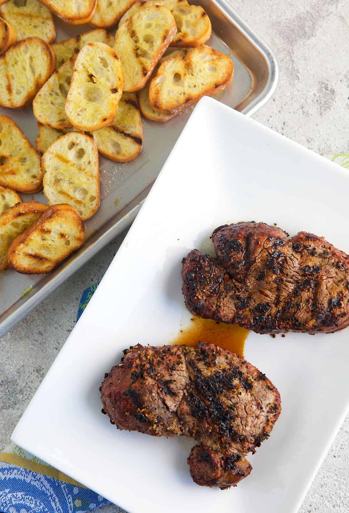 Two cooked steaks are placed next to a sheet pan filled with crostini.
