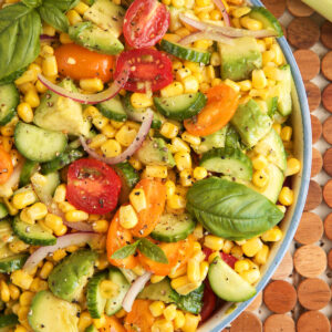 Overhead view of avocado corn salad in bowl with tomatoes, cucumbers, and fresh basil