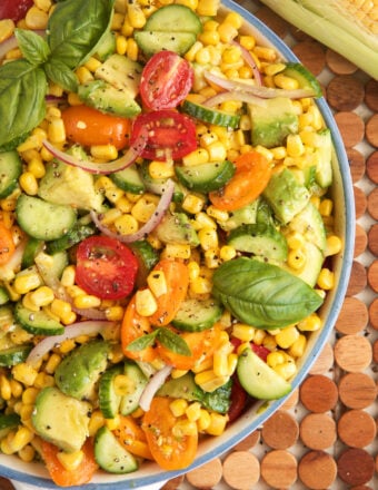 Overhead view of avocado corn salad in bowl with tomatoes, cucumbers, and fresh basil